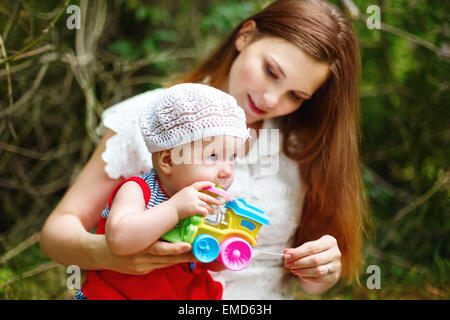 Cute Toddler Baby Girl sitting on Mom`s Hands, playing with Toy. Relaxing in green summer Park. Selective focus on baby`s eyes. Stock Photo